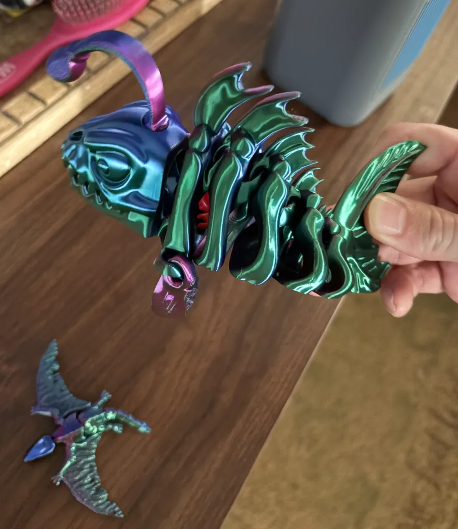 Close-up of an articulated skeleton angler fish 3D print held in a hand, its color-shift filament showing green and blue with a red interior detail