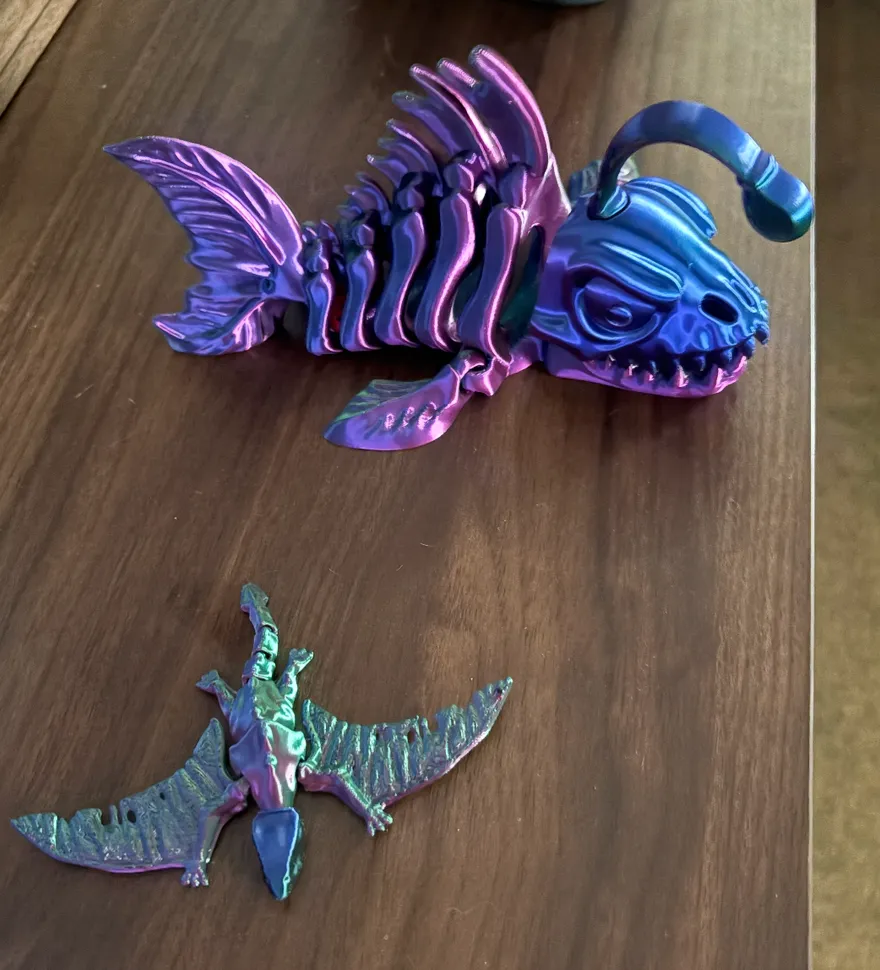 Two articulated 3D prints on a wooden table: a skeleton angler fish and a pterodactyl, both in color-shift filament that fades between purple, blue, and green
